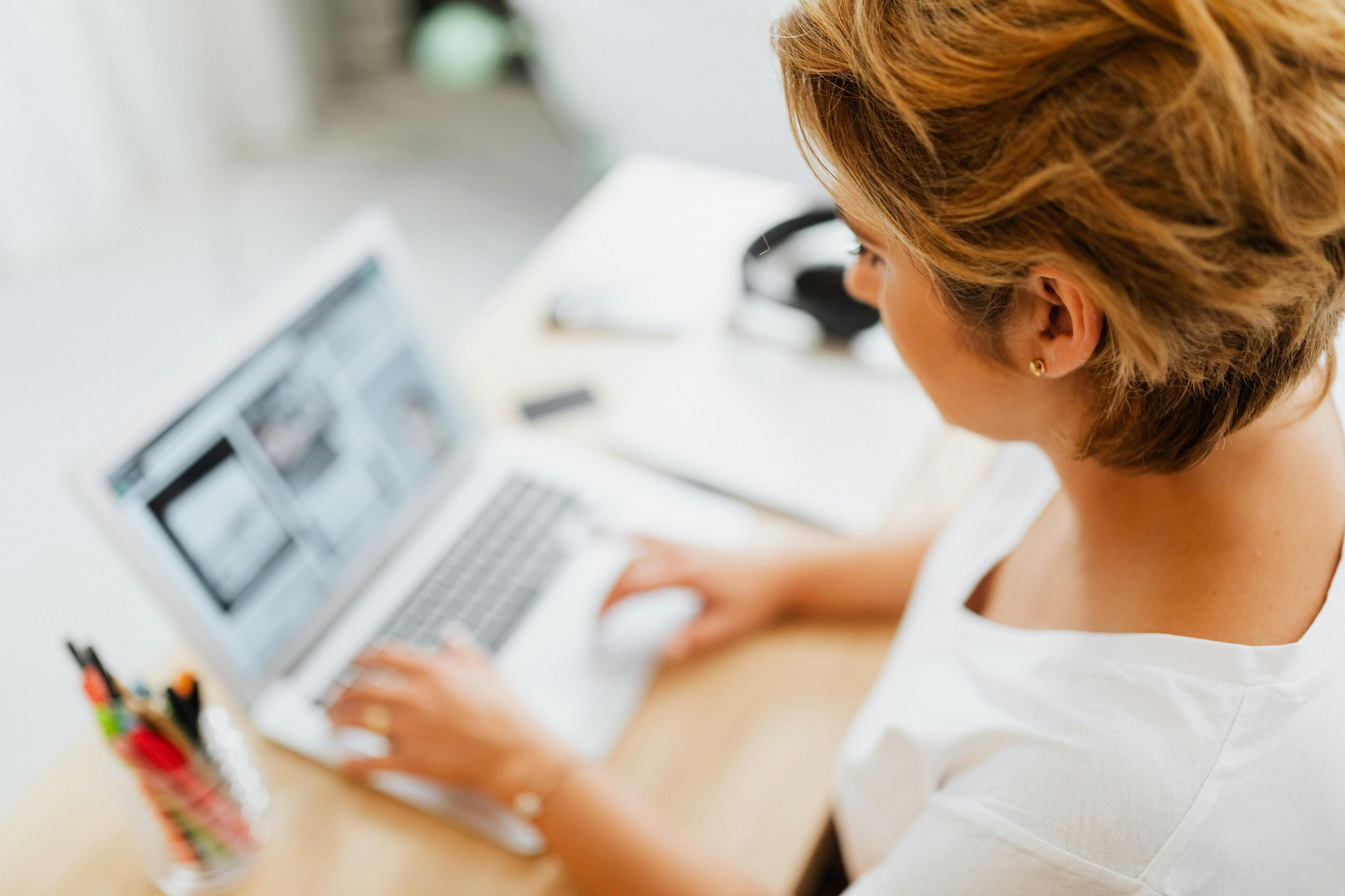 Frau mit blondem Haarknoten arbeitet konzentriert am Laptop in hellem Büro mit verschwommenem Hintergrund und Stiftebecher auf dem Schreibtisch.