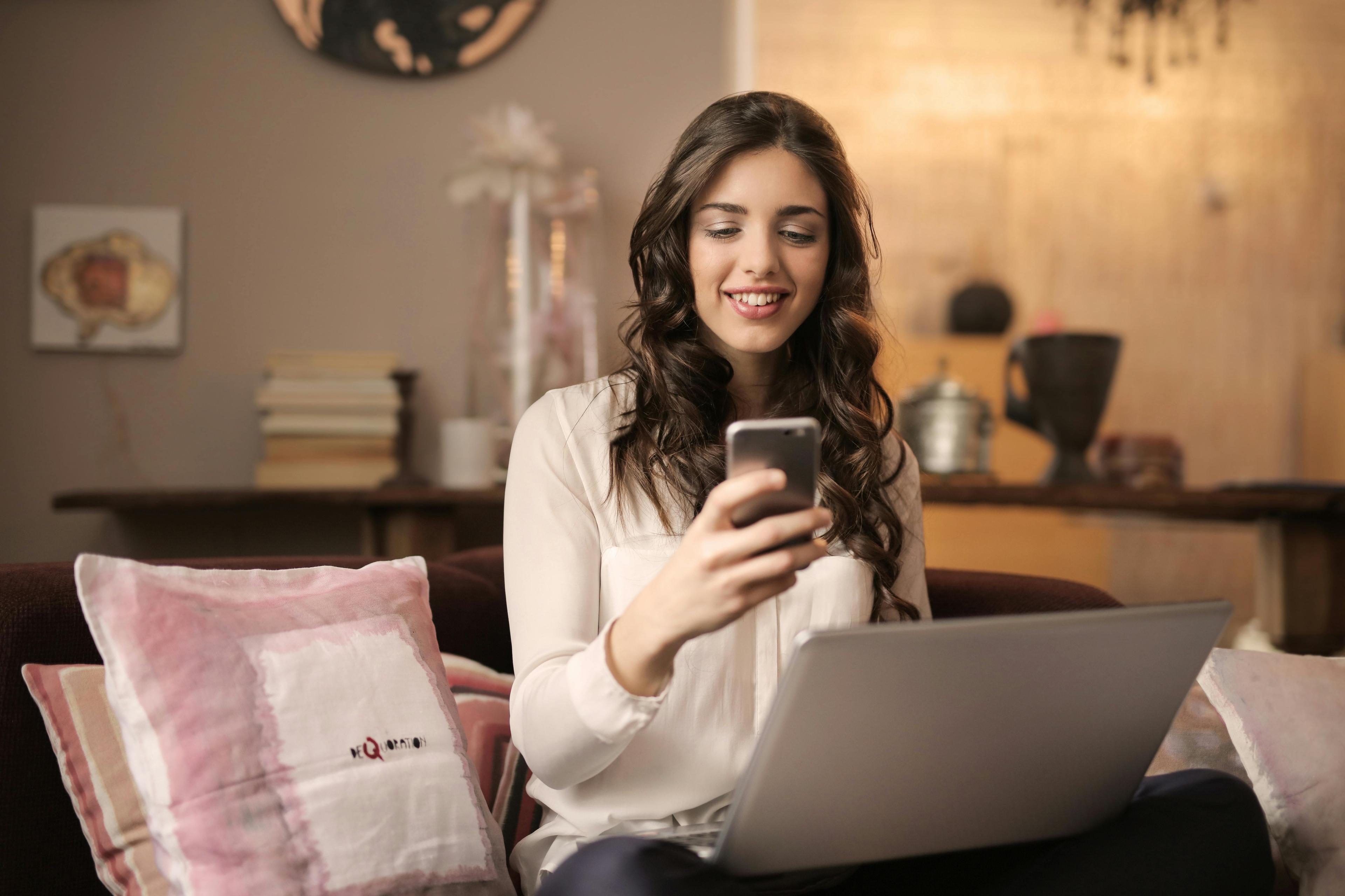 Lächelnde junge Frau mit langen dunklen Haaren in weißem Pullover nutzt Smartphone und Laptop gleichzeitig auf Sofa mit rosa Kissen in gemütlichem Wohnzimmer.